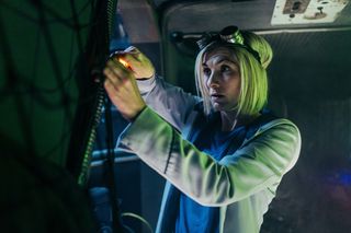 The Doctor (Jodie Whittaker) is doing repair work on some cabling in a darkened room, using her sonic screwdriver. She is wearing goggles, pushed back on top of her head.