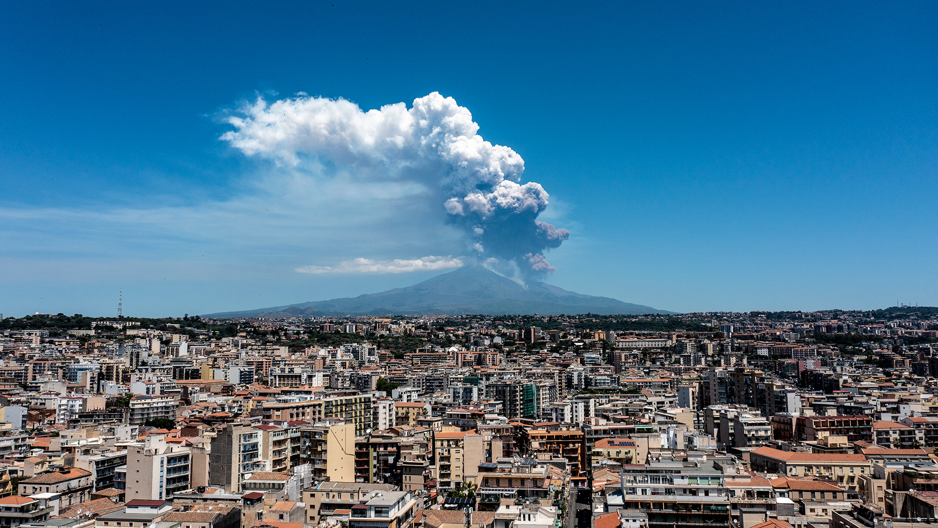 A volcano erupts in the background behind a busy town