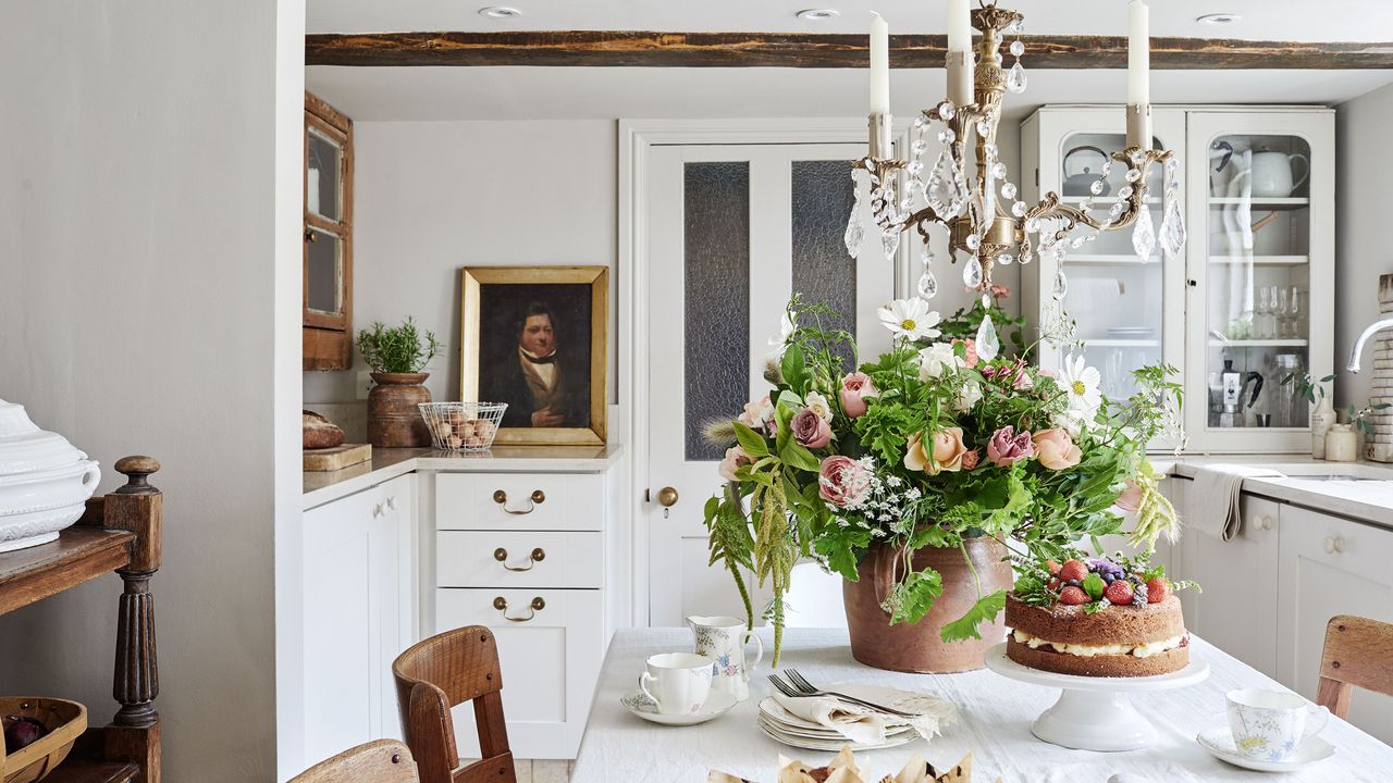 kitchen with white tablecloth and white cabinets and walls and candle chandelier with Real Flower Company bouquet and cake and sink and part-glazed door in background