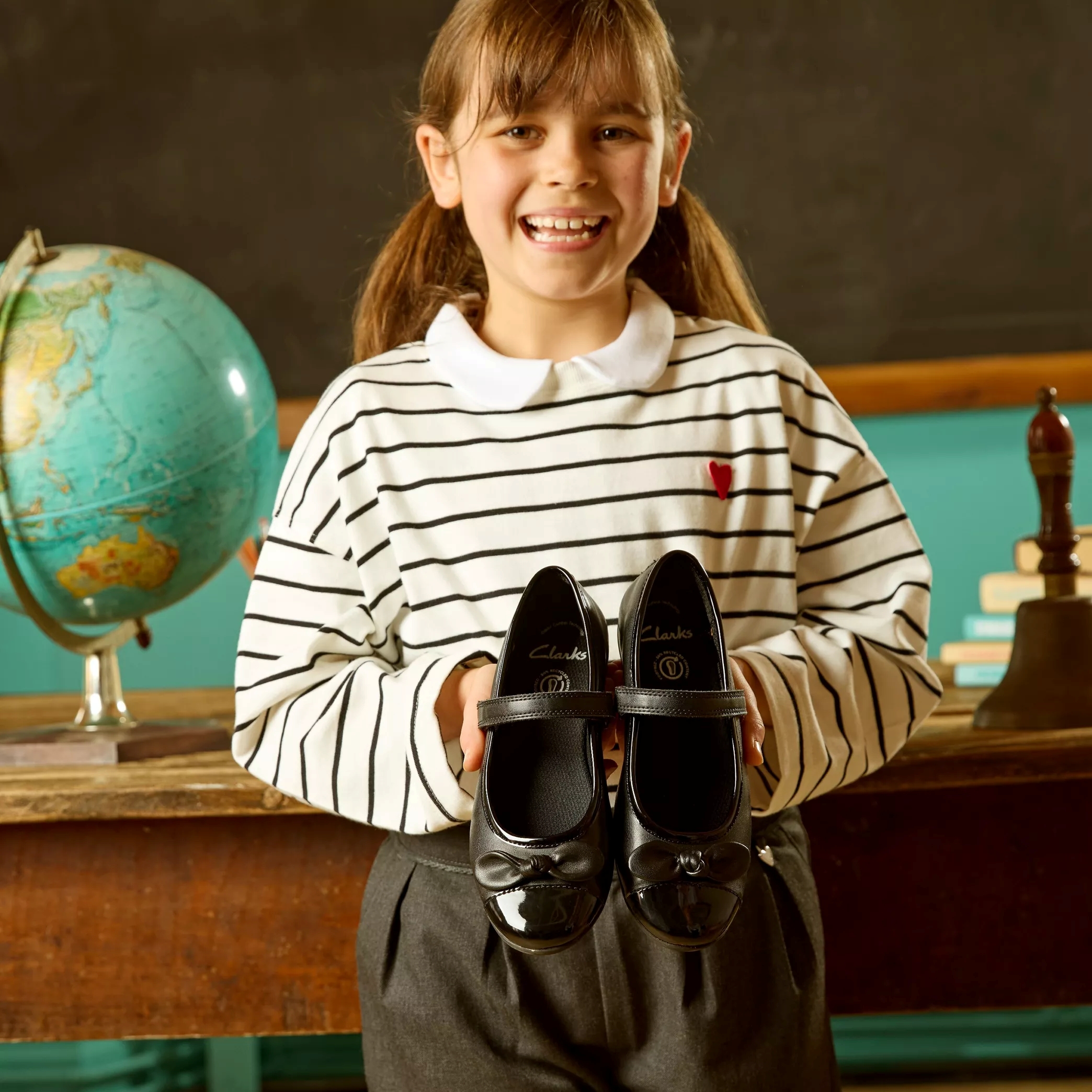 Child holding black mary jane school shoes sold at Clarks in front of a desk with a globe on it 