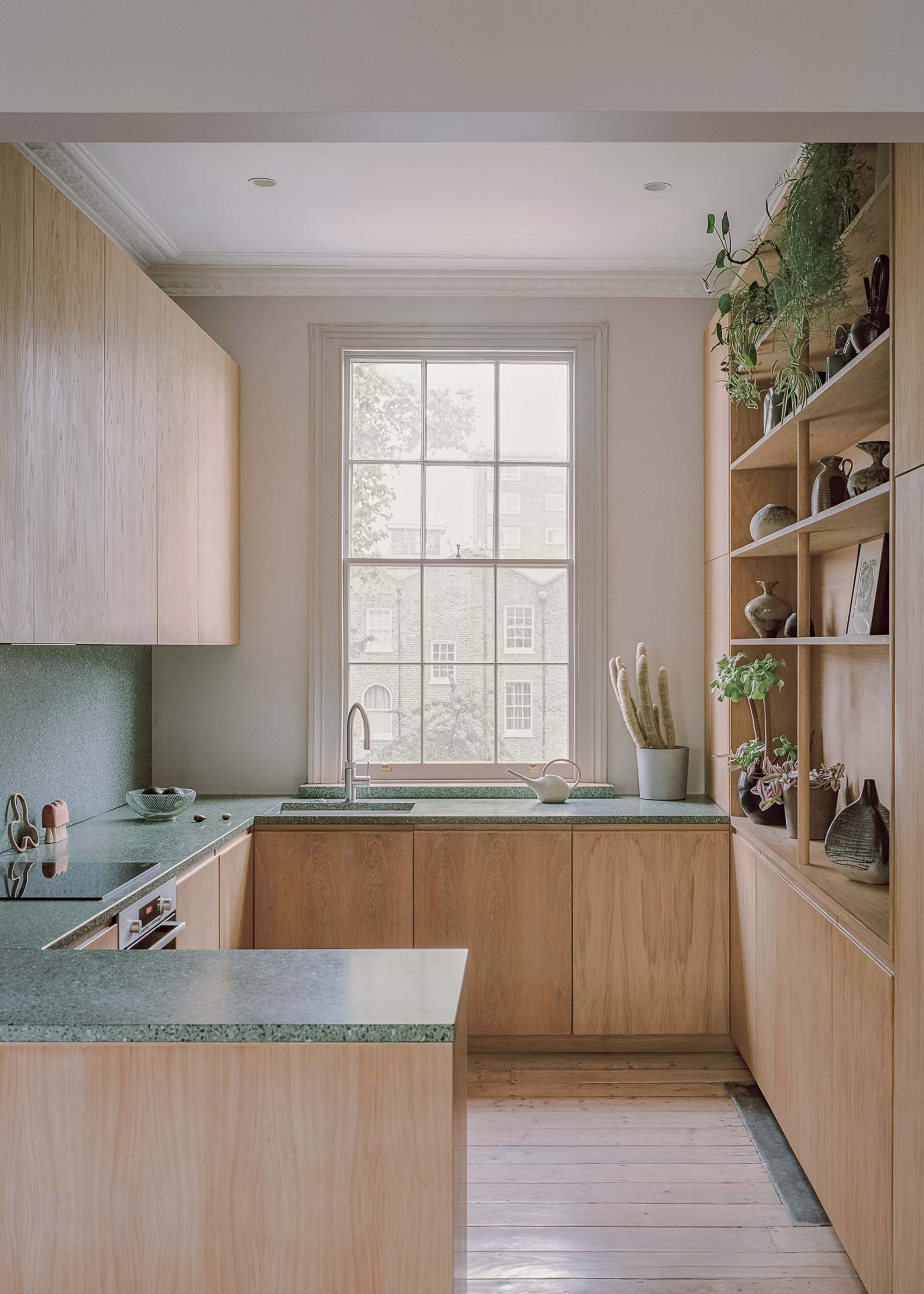tiny kitchen with light oak cabinetry and dark green terrazzo counters, and open shelving with plants and pottery on one wall