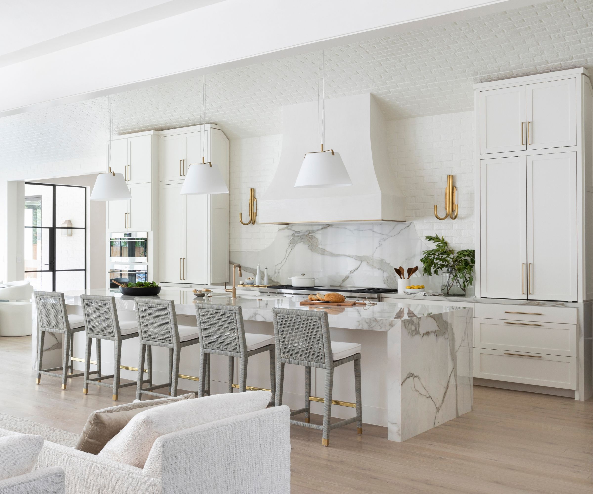 A white open plan kitchen with marble counters, a plaster range hood, and a layered lighting scheme