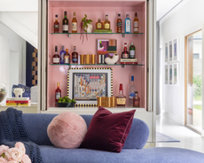 A living room with a blue sofa, and behind that a divider wall containing a pink-toned bar unit