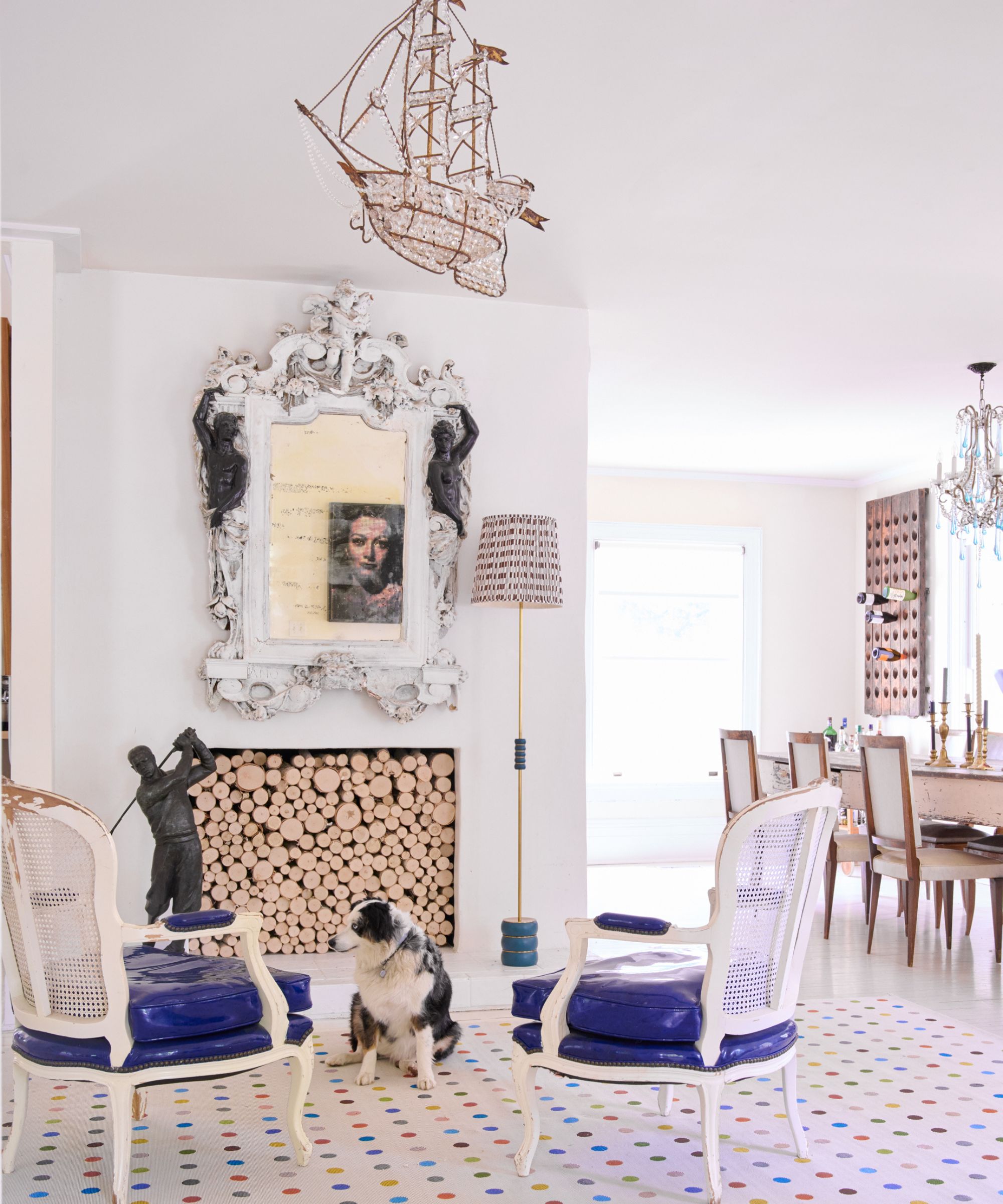 white dining room with antique mirror above the fireplace which is filled with logs and surrounded by traditional white chairs on to a modern spotted rug
