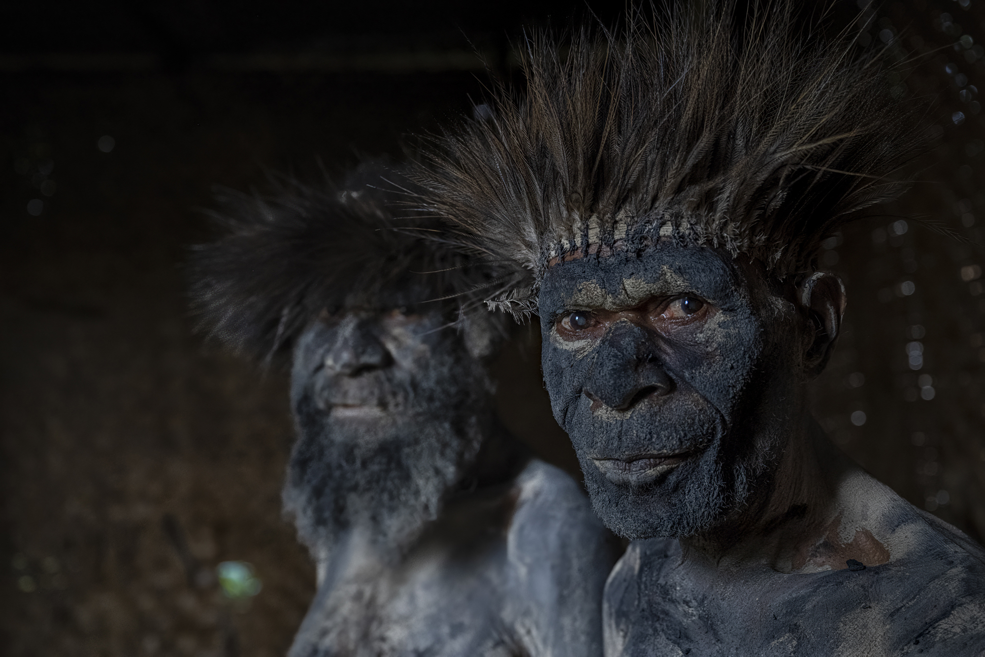 Two Wauga elders stand together, among the very few who remain as guardians of their tribe’s traditions. Their weathered faces, marked by deep wrinkles, reflect a lifetime of wisdom and resilience. Adorned with intricate feathered headdresses and covered in black mud, they represent a culture at risk of fading. The elder in the foreground gazes thoughtfully, bearing the responsibility of preserving their legacy. Behind him, the second elder mirrors the same quiet strength, their shared presence a symbol of the community’s enduring bonds. This powerful image captures the fragility of their cultural rituals, reminding us of the importance of safeguarding traditions before they are lost forever