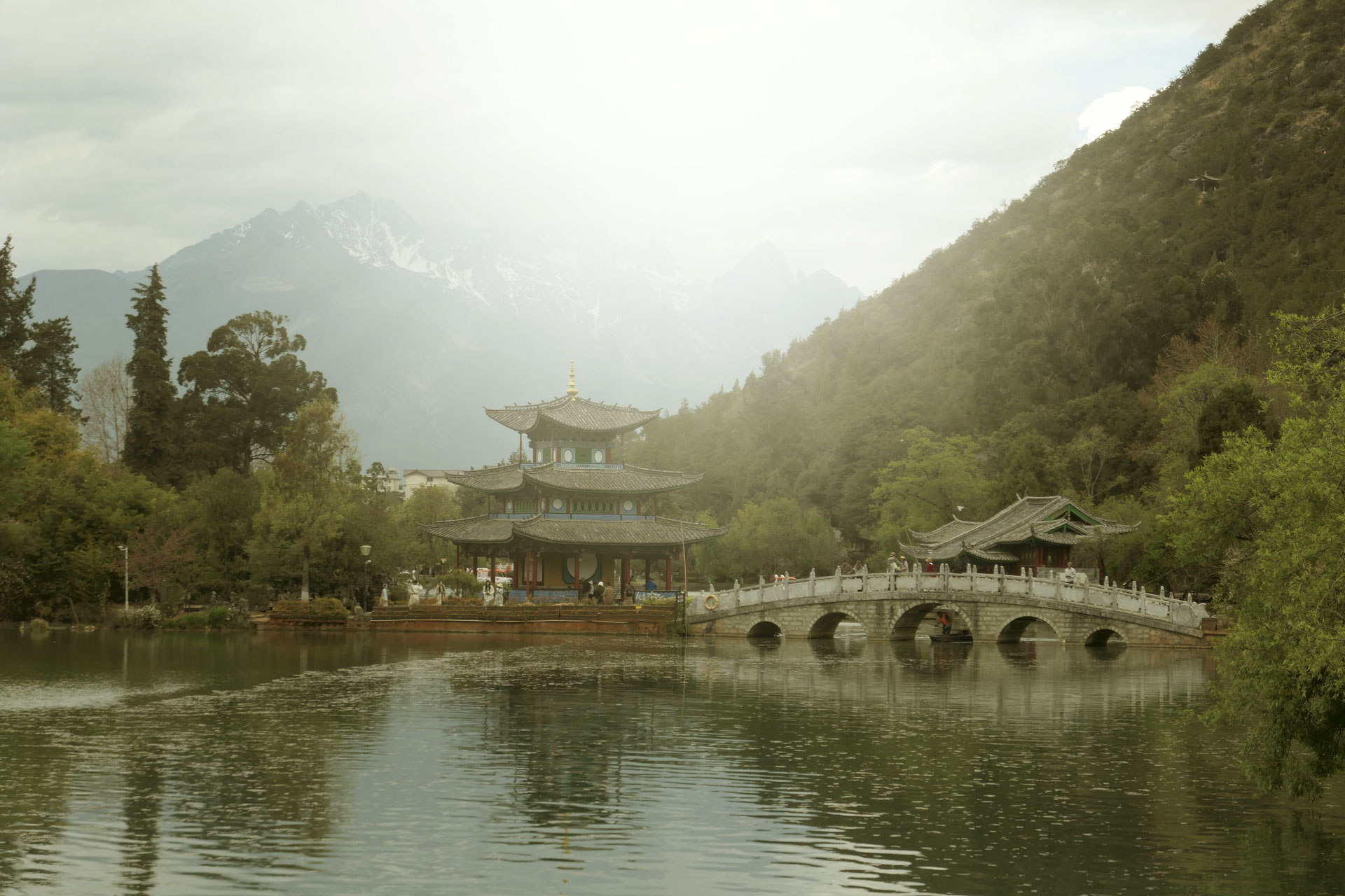 A Chinese temple by the side of a lake with an arched bridge leading up to it and both are reflected in the still water, in front of a snow covered mountain. A creative filter is applied to the image