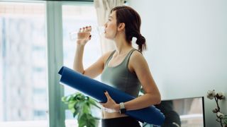 woman holding a yoga mat, refreshing with water after practicing yoga at home