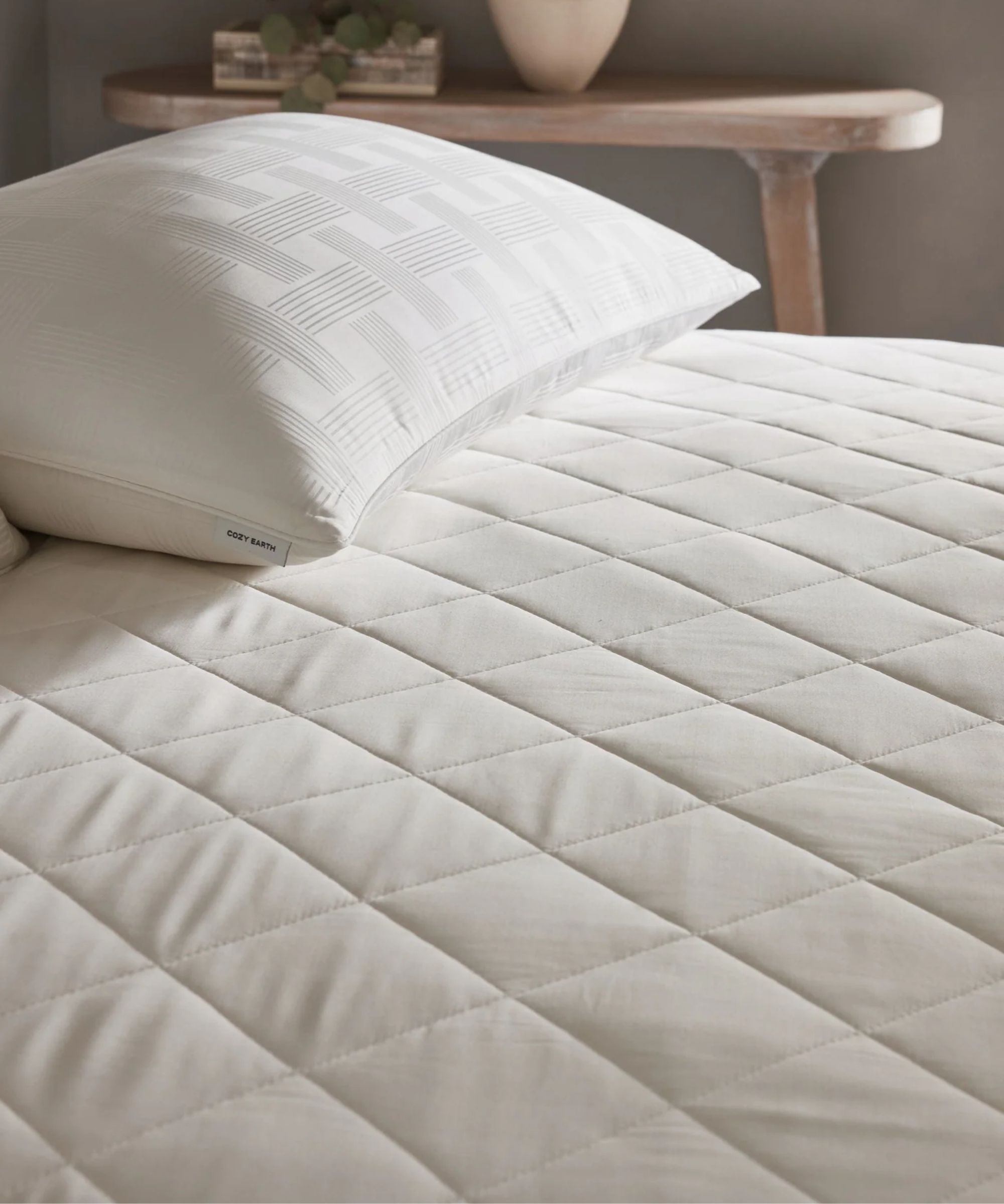 A white quilted Cozy Earth Bamboo Mattress Pad shown on a bed, with a white pillow to the top right of the bed. Behind a wooden side-table is seen, with a green houseplant in a brown rectangular pot, and a white plant pot beside it.