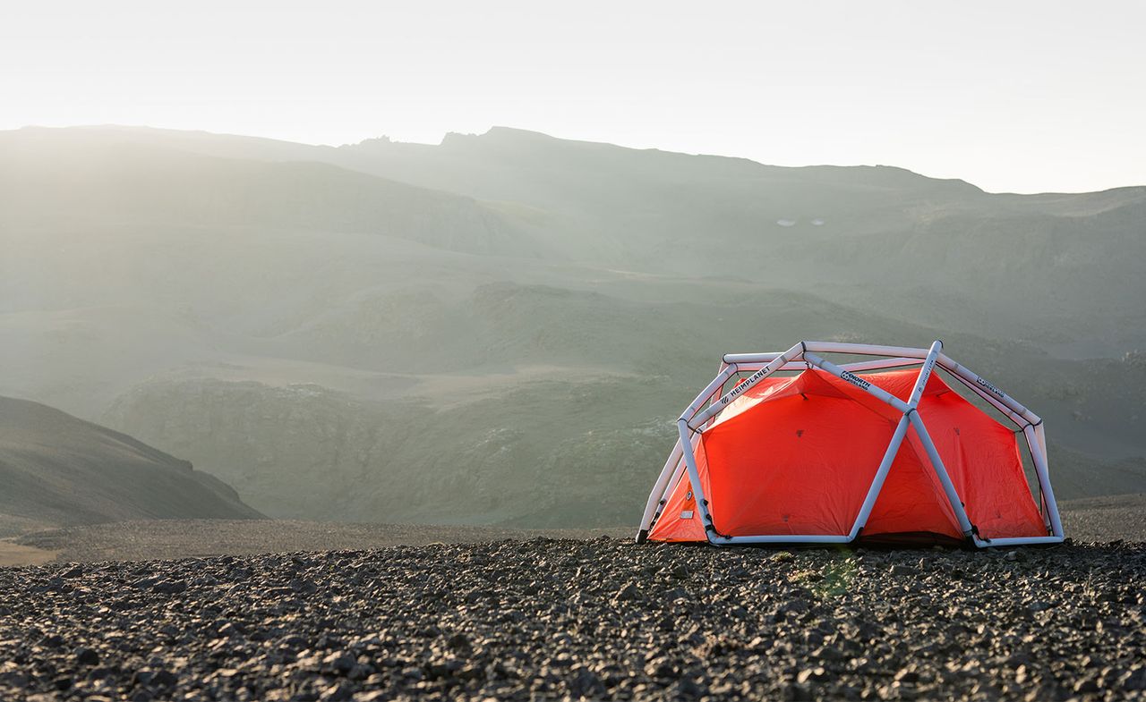 View of an orange tent with a white framework by 66°North and Heimplanet during the day - the tent is pictured on rocky ground outside close to some hills