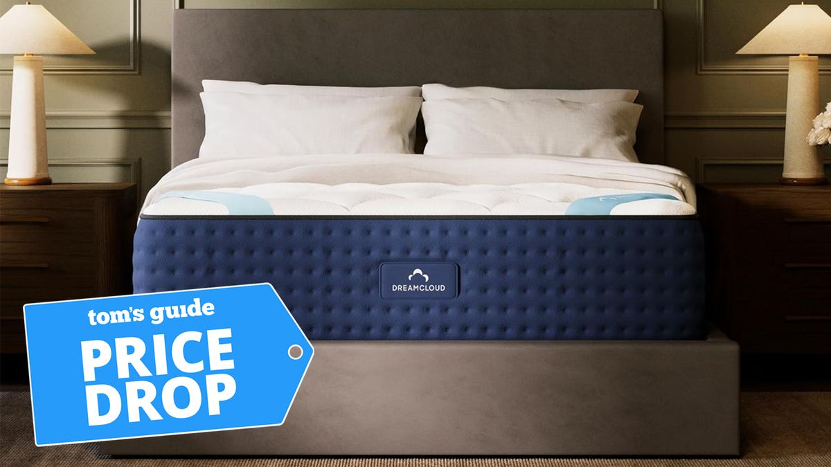 The DreamCloud mattress shown on a grey fabric bed frame with a blue price drop sales badge overlaid on the image