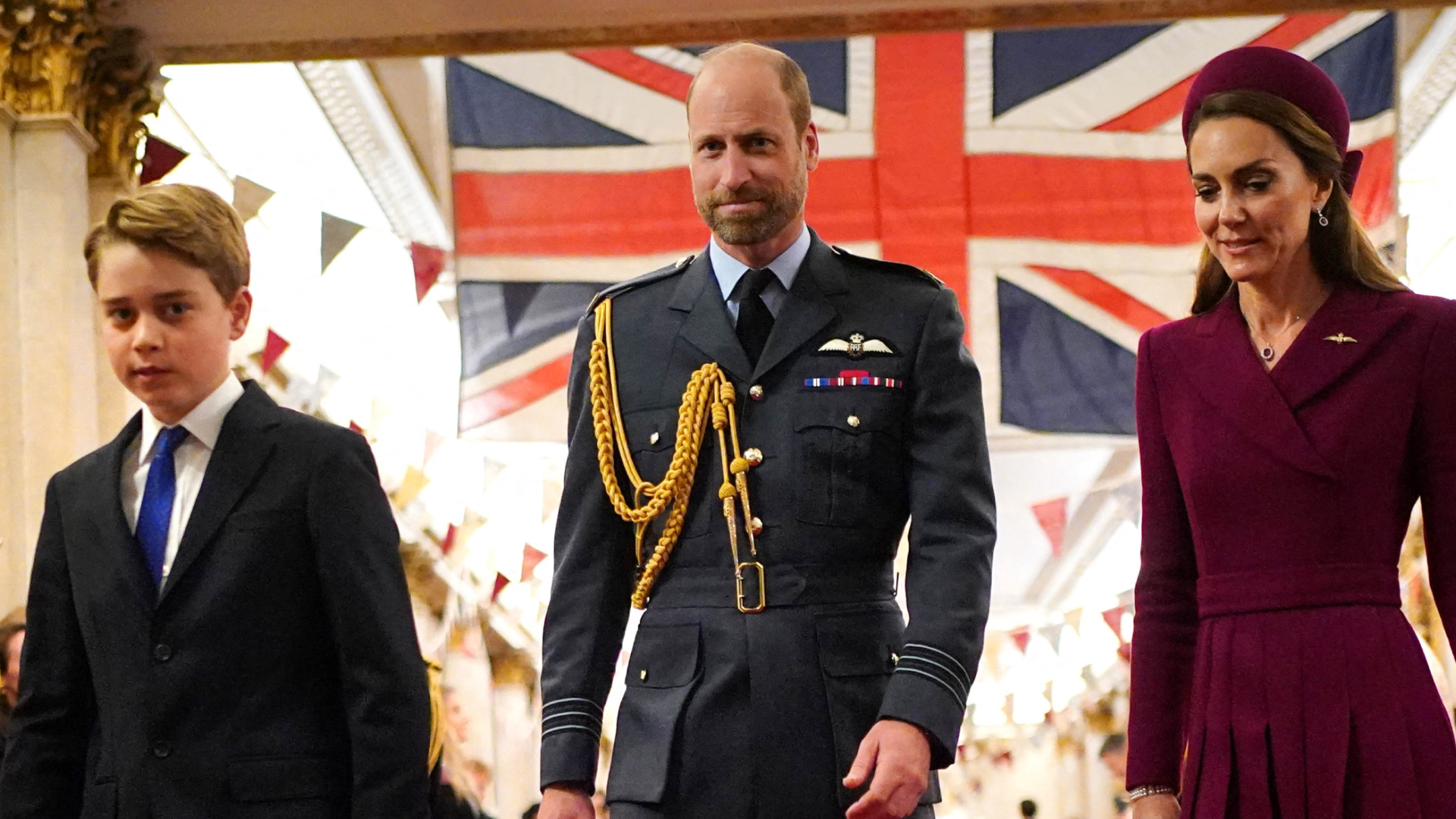 Prince George wears a suit, Prince William wears military uniform, Kate Middleton wears a dark red coat dress, as they walk in front of a Union Jack flag