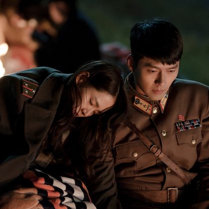 a woman wears a military jacket and rests her head on a soldier's shoulder outside of a fire in a still from crash landing on you