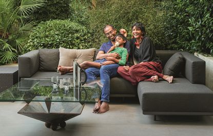 a family sat on an outdoor sofa in front of a lush green backdrop