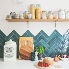 A kitchen work top area with green tiles and white surface. On top are three chopping board stacked against the wall, a vintage bread flour jar, salt and pepper mills, basil plant, salt pot, bowl of onions and board with fresh bread and tomatoes on top.