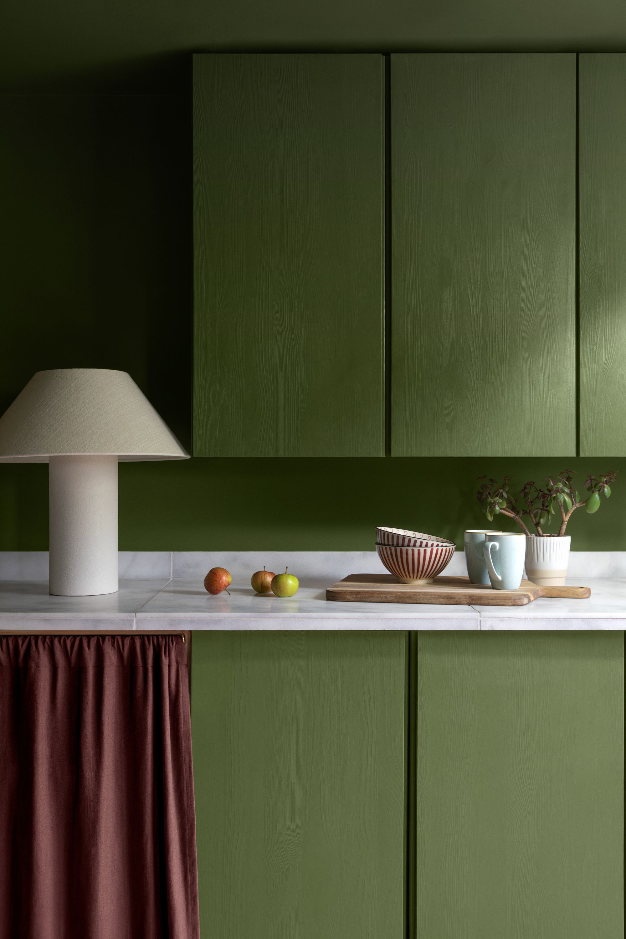 Kitchen cabinets painted in a bold green with a red cabinet curtain and a white fabric lamp on the counter.
