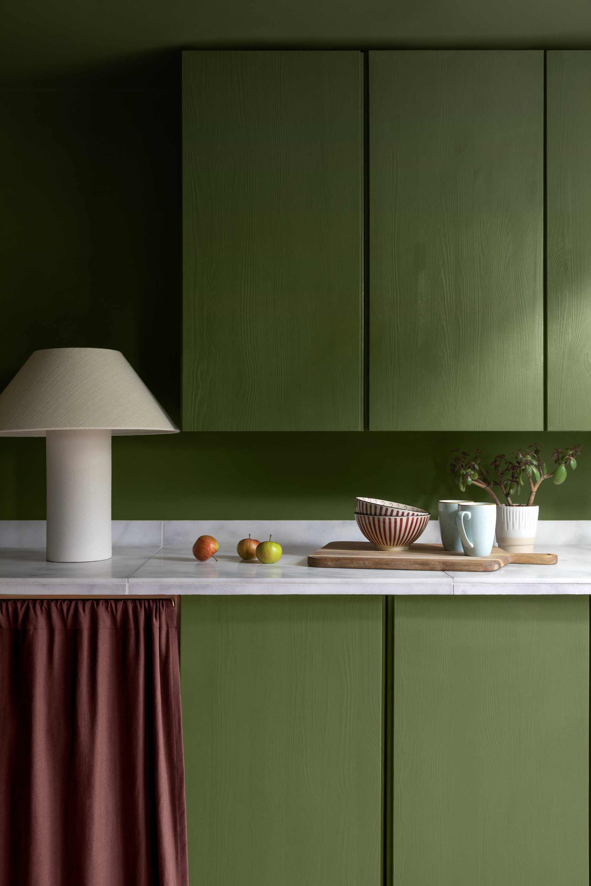Kitchen cabinets painted in a bold green with a red cabinet curtain and a white fabric lamp on the counter.