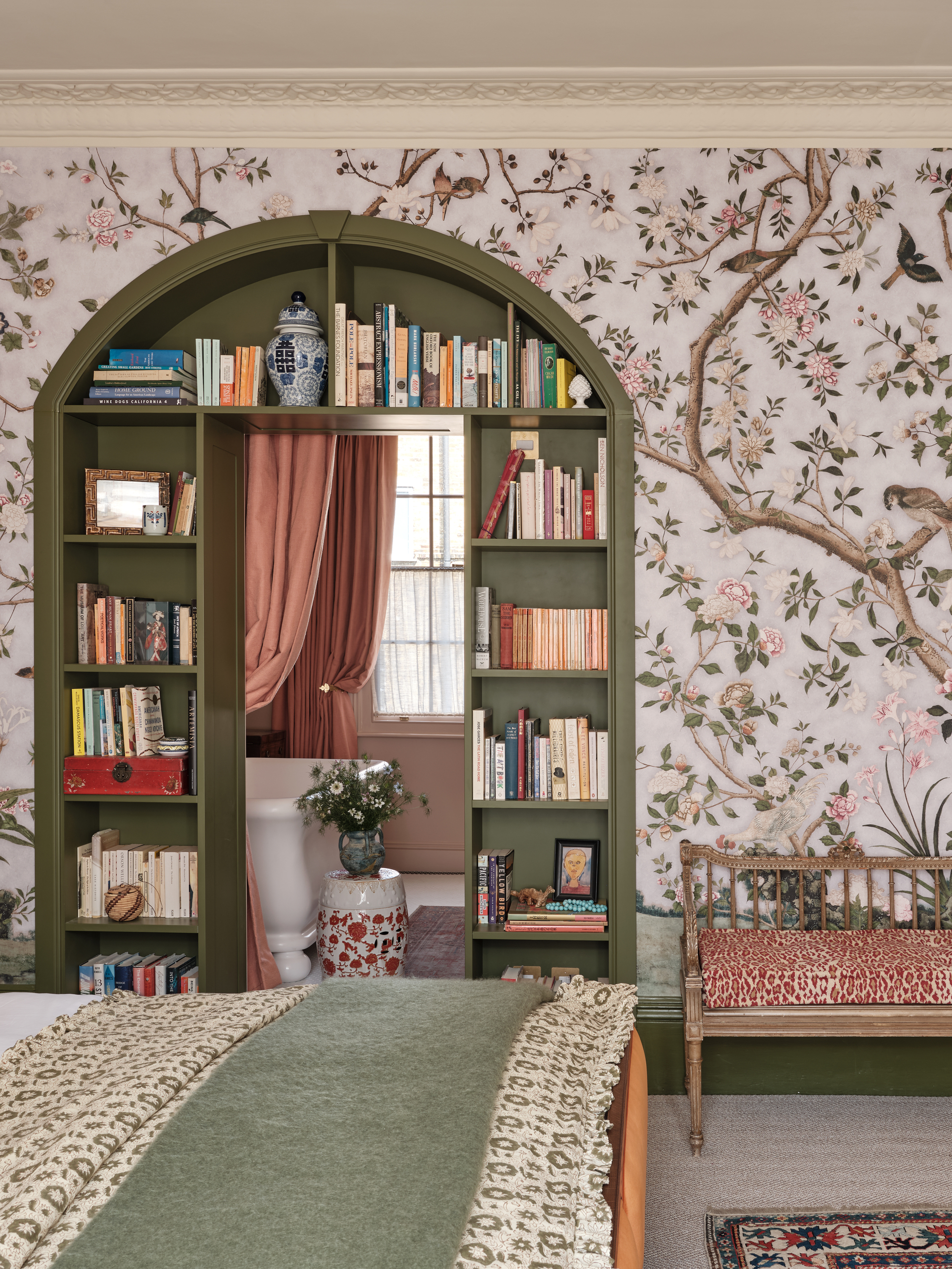 A bedroom with a chintzy patterned wallpaper and a built-in bookshelf around the doorway