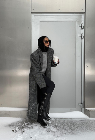 what to wear in 30-degree temperatures, shown in a photo of a woman standing outside in the snow wearing a black balaclava with a long herringbone gray coat, gray crewneck sweater, thermal leggings, and black moto boots