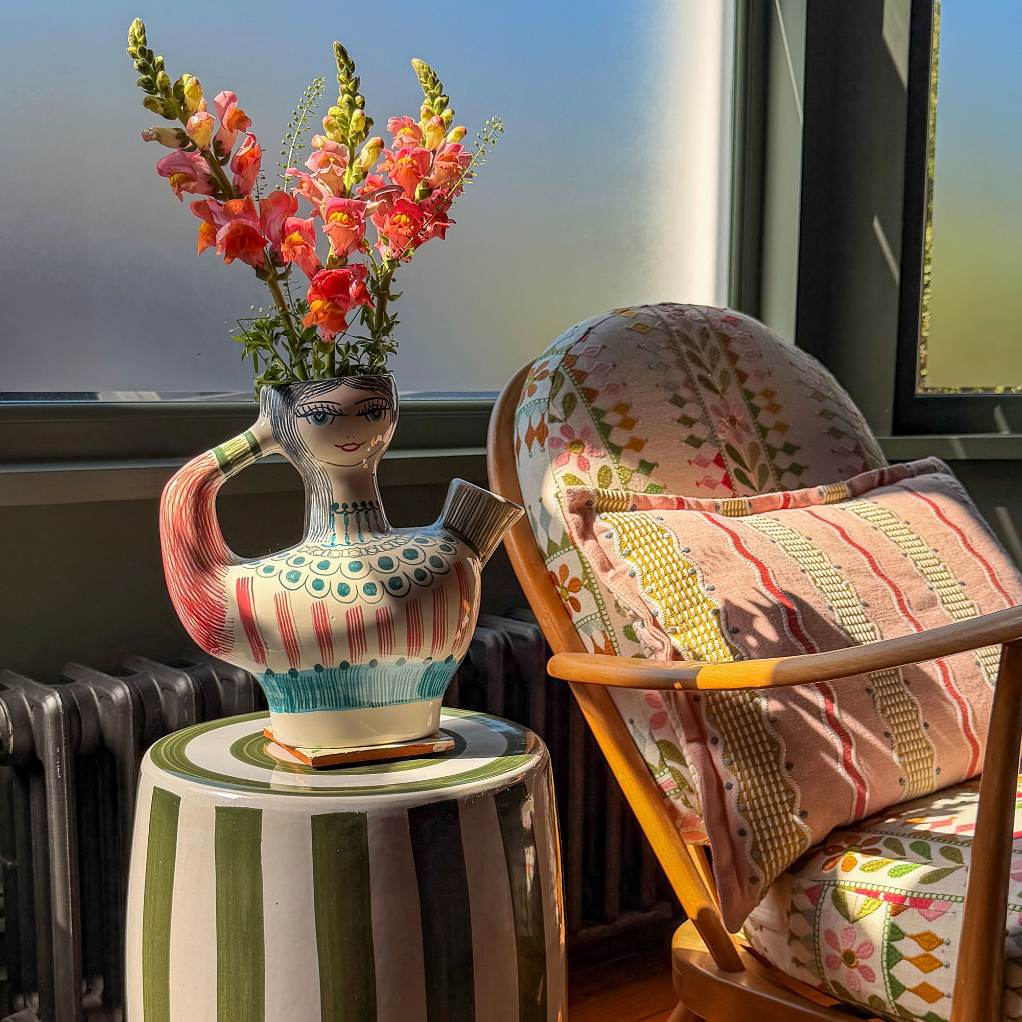 Woman-shaped vase filled with flowers sitting on green and white striped ceramic stool beside wooden armchair with colourful patterned upholstery and cushion