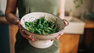 A woman in a khaki green sleeveless top holds a white colander full of freshly washed raw spinach, a magnesium rich food that has multiple benefits to a person's health and sleep