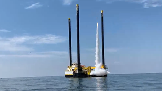 a rocket lifts off from a floating platform in the sea. blue sky is behind. three small launch towers surround the platform