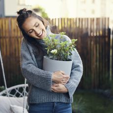 A smiling woman in a sweater holds a houseplant in her backyard
