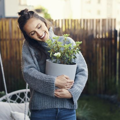 A smiling woman in a sweater holds a houseplant in her backyard