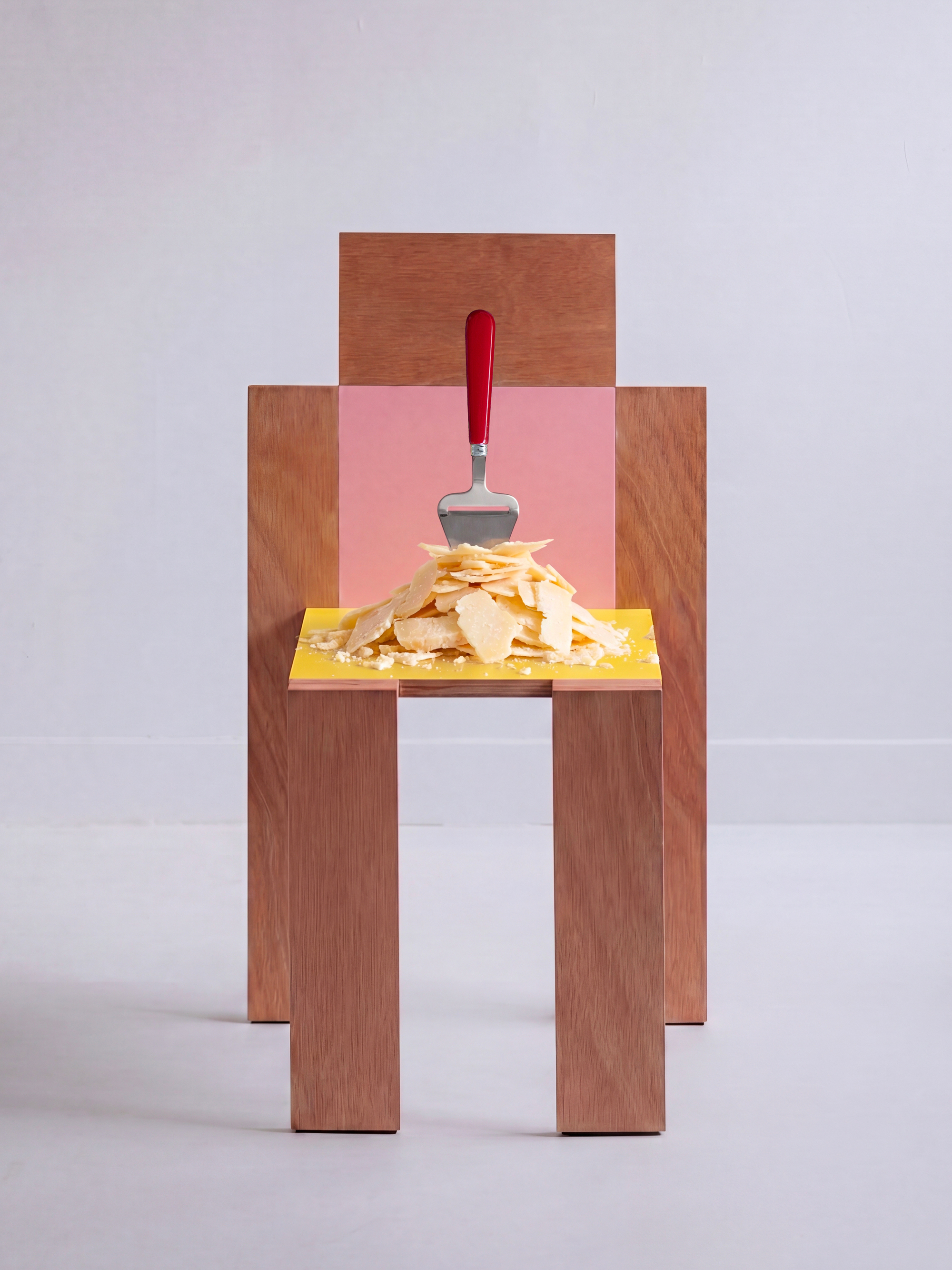 Image of a red cheese slicer stuck into a pile of sliced cheese on a wooden chair with a yellow top and a pink back in an all-white room.