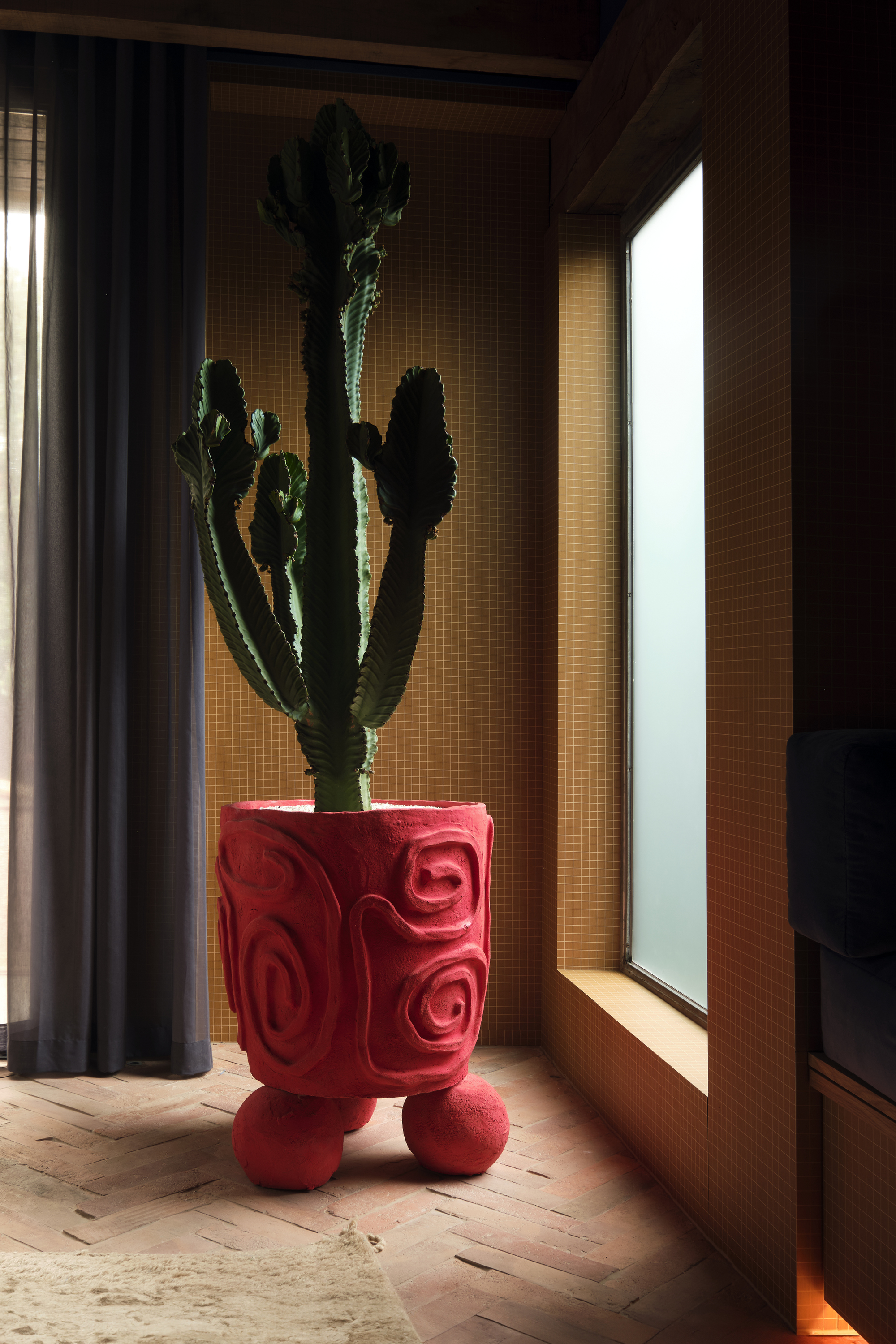 A large cactus plant in a hand-made, bright red, large vase with ball feet. The vase is in the corner of a room with brick flooring and yellow subway tile walls with a large window.