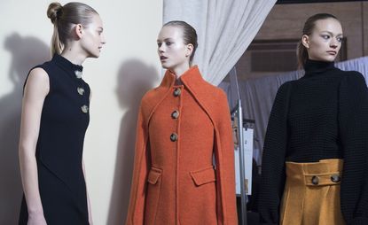 3 Models wearing black full length sleeveless dress, orange coat, black jersey and mustard skirt