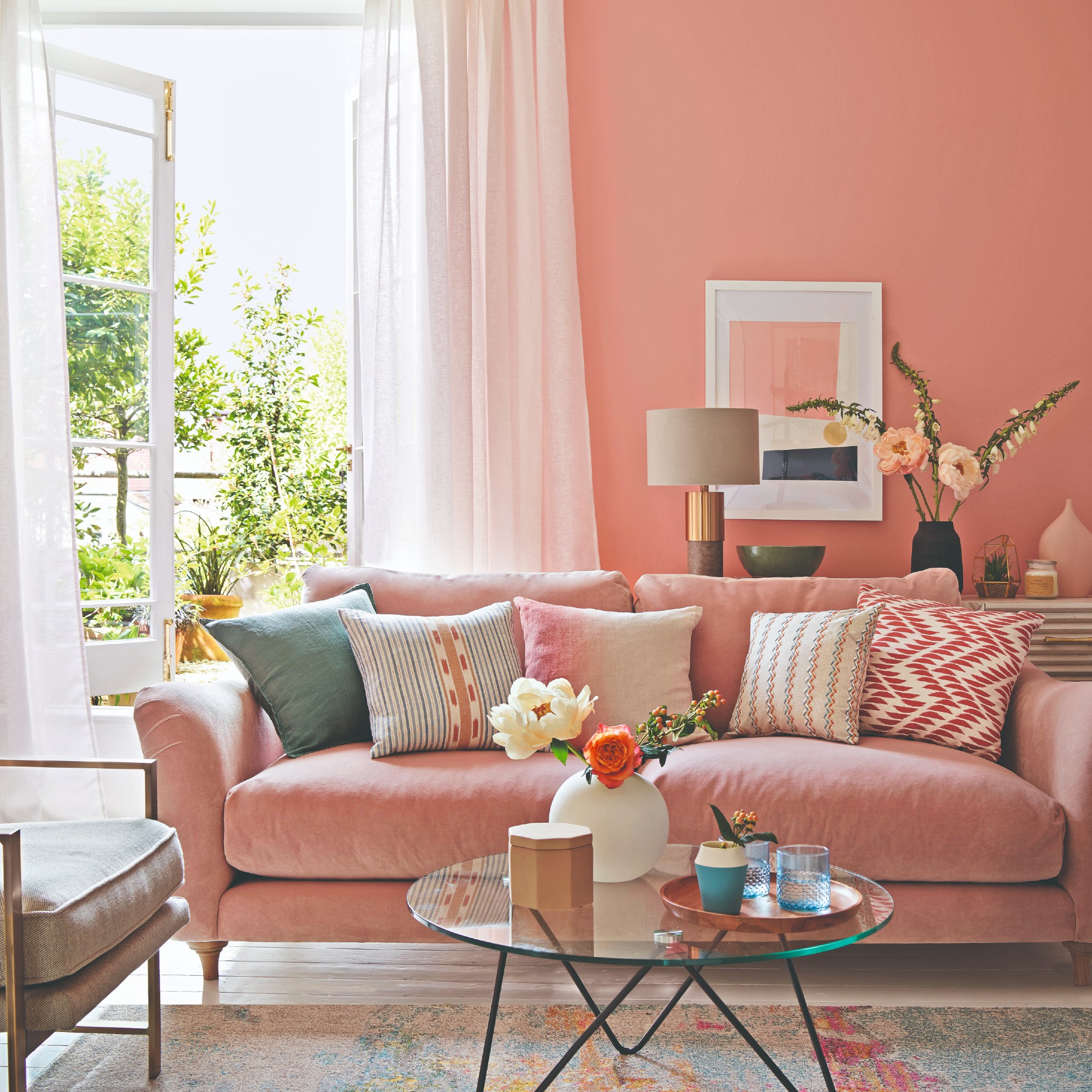 A peach-coloured living room with a tonal velvet sofa with a large window to the garden framed with floor-length voile curtains