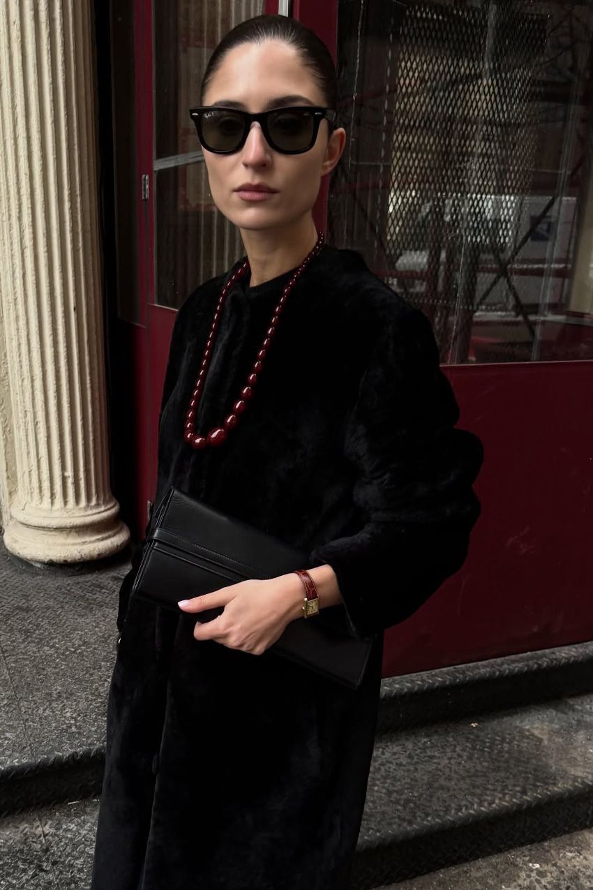 A woman standing outside in front of a red storefront wearing a long black pony-hair coat with a long red resin bead necklace, a dainty red watch, a black clutch bag, and black Ray-Ban sunglasses.
