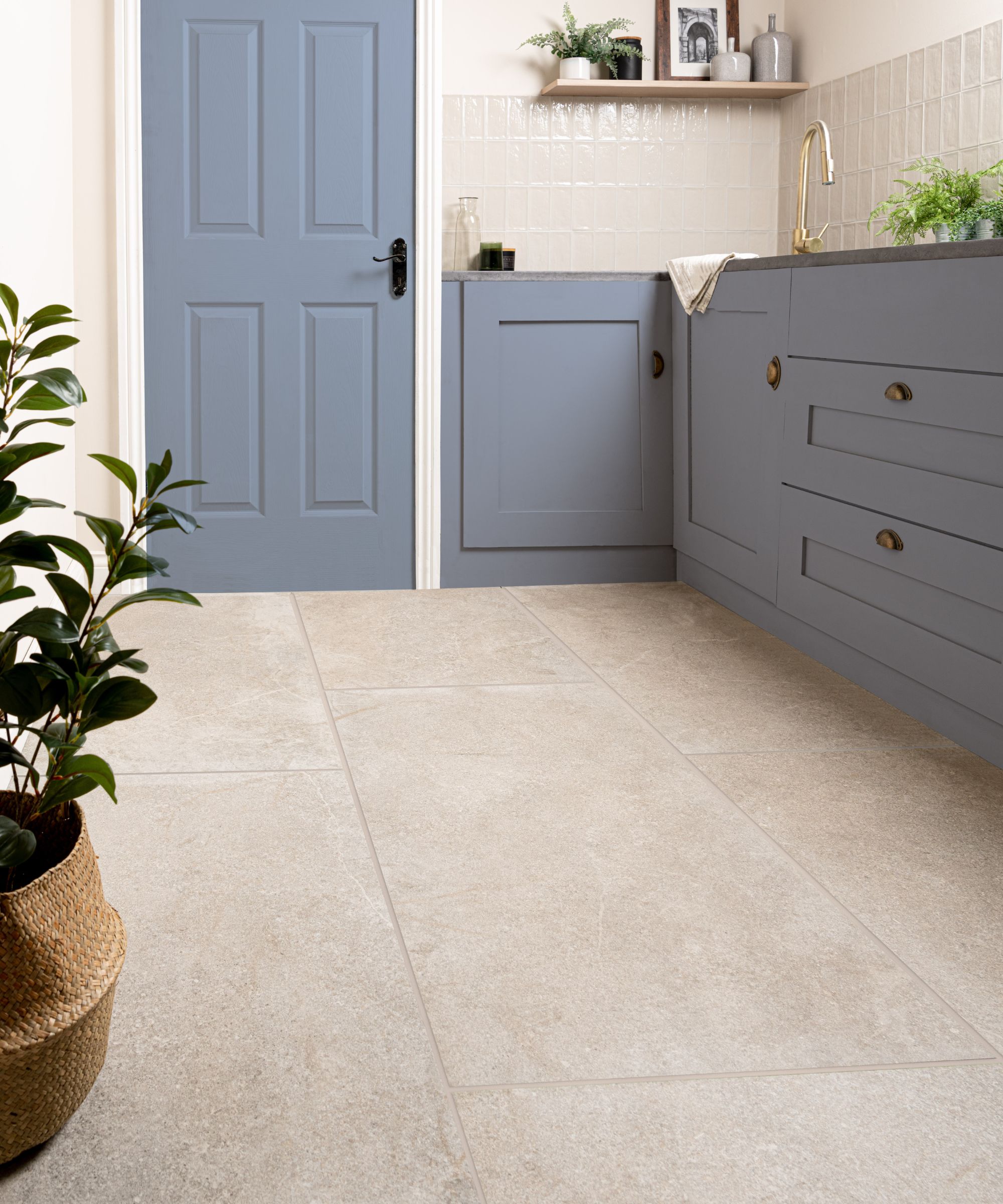 Modern utility or laundry room with soft beige stone-look floor tiles, blue cabinetry with brass hardware, a blue door, and neutral wall tiles. Green potted plants add a fresh touch.