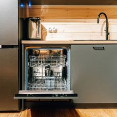 Open integrated dishwasher next to a fridge freezer, in a kitchen with wood panelling, a wood-look worktop, and sage green cabinet doors and black handles