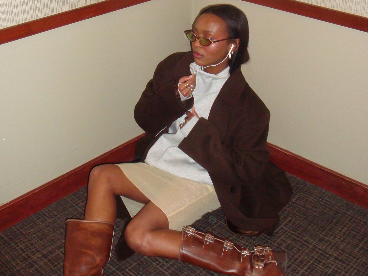 Devon Apollon wearing a brown jacket, gray zip-up hoodie, khaki skirt, and brown buckle boots.
