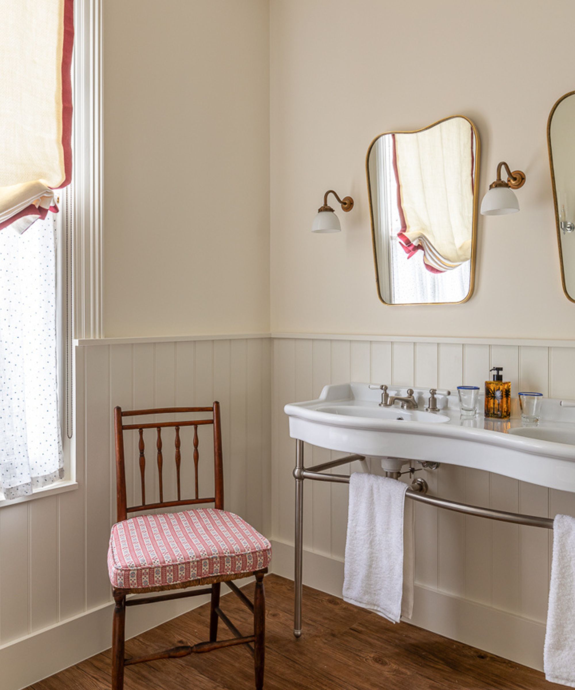 Bathroom with wood panelling, cream walls, wooden floor, mr and mrs sink, gold sconces flanking curved mirrors, and a red vintage chair