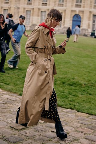 a woman at Paris Fashion Week in a khaki trench coat, black and yellow polka dot dress, and heeled black boots