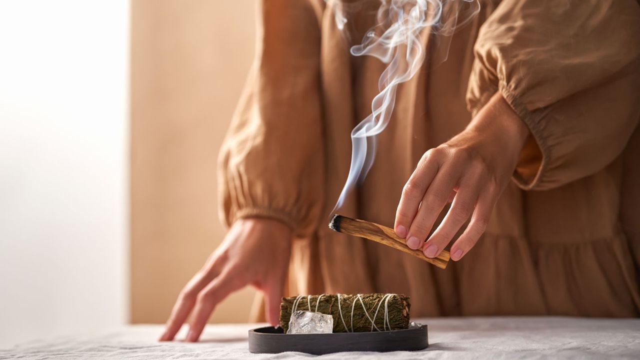 The hands of a girl in a beige dress hold a smoking palo Santo stick against the background of ritual things: a rock crystal stone and a twist for fumigation.