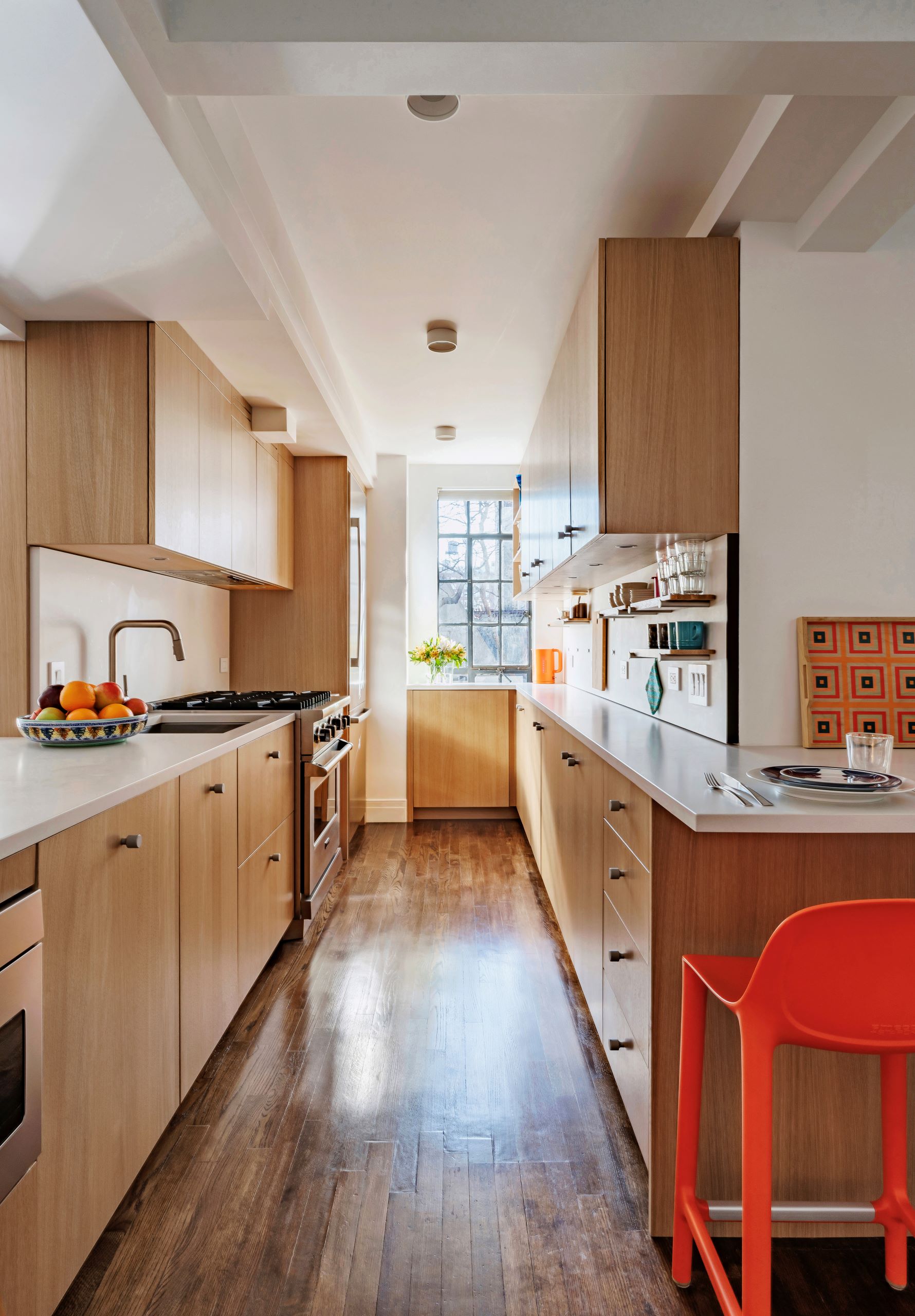A galley kitchen with wooden cabinets, and a countertop with orange bar chairs