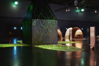 A black pentagon shaped sculpture lit up with projections of green stick like designs in a dark room of a museum gallery