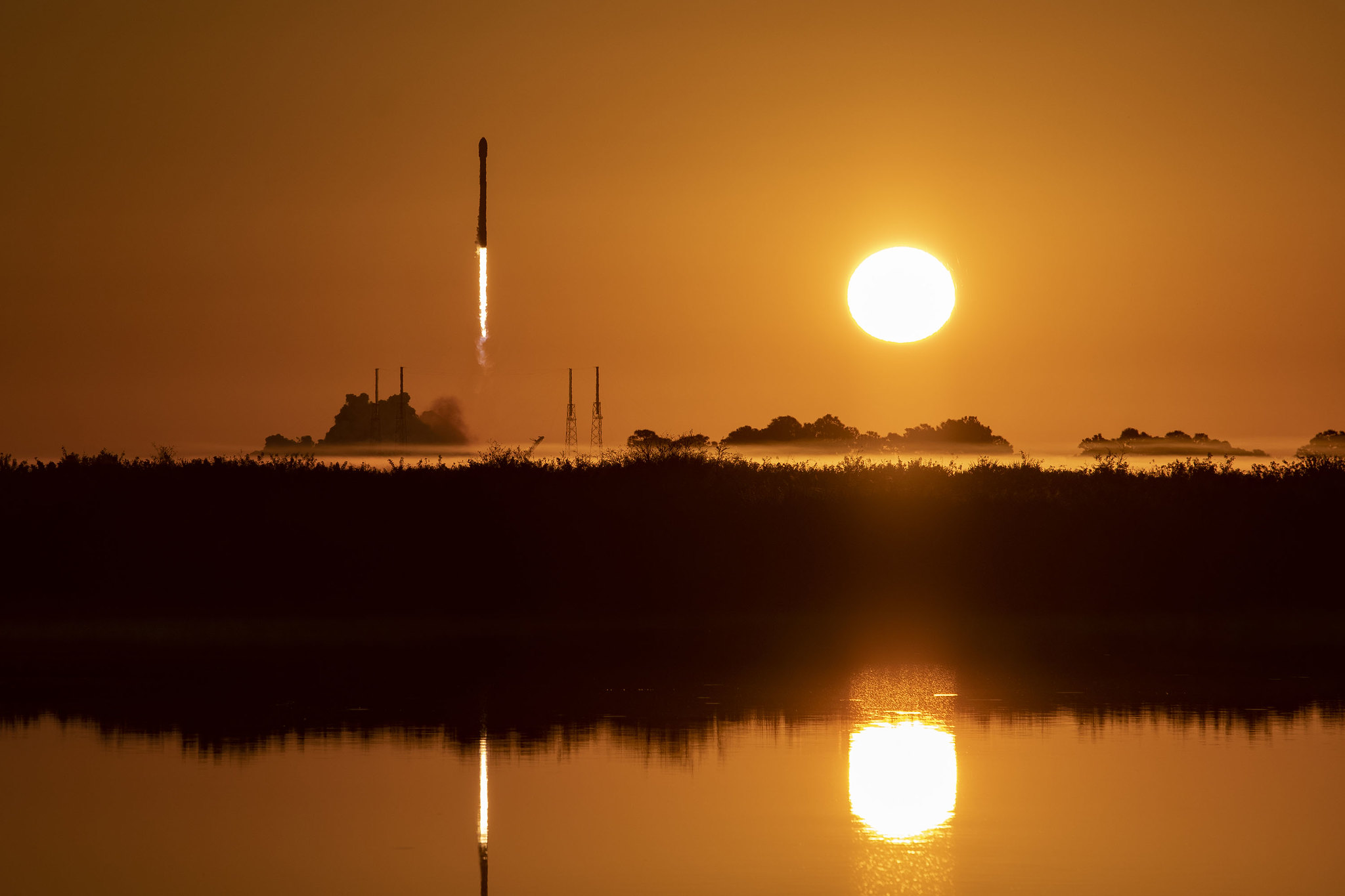 a spacex rocket launching with the rising sun beside it, reflected in water