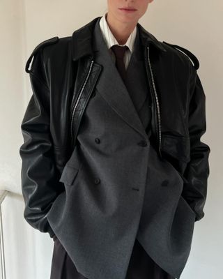 A photo of a woman wearing a gray double-breasted Saint Laurent S/S 25 suit with a striped shirt, tie, and leather bomber jacket.