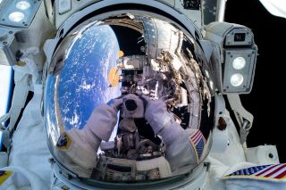 the mirrored visor of a space suit shows the reflection of earth and two hands holding a camera.