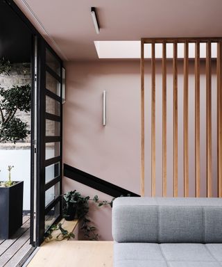 colors that go with light pink, light pink living space with light grey couch. Wooden slats near staircase, view of garden, black crittall doors, tree in pot, pale wood floor, skylight