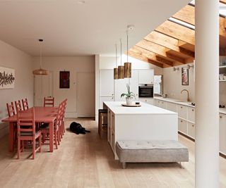 inside of contemporary home with timber beams to glass roofed side extension, white kitchen, wooden floor and pale terracotta dining set