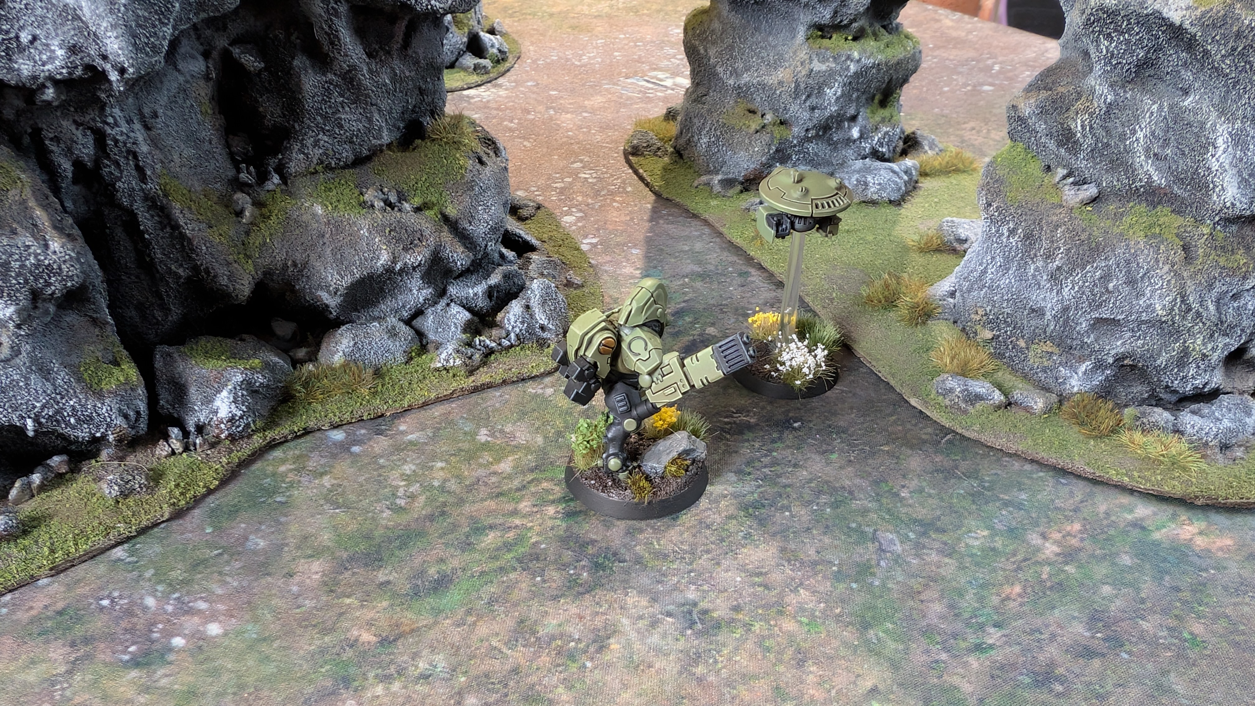 A Tau Stealth Battlesuit model and drone navigate through towering rock formations on a grassy battlefield