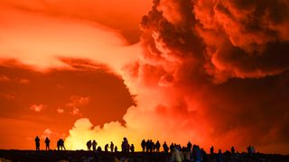 a large cloud of gas is illuminated at night by glowing lava as silhouetted bystanders look on