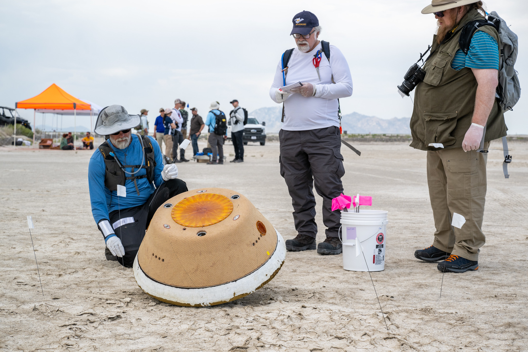 several people stand in the desert looking down at a cone-shaped capsule on the ground