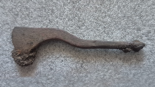 an oxidized iron tool with one spatulate end and one pointed end rests on a grey speckled table