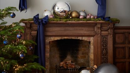 A fireplace with the mantel decorated with metallic oversized baubles and a tree next to it